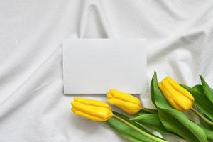 three yellow tulips lie on a draped linen bedspread with a white sheet of paper photo