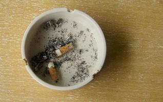 Two cigarettes on white ashtray with ashes inside it. Wooden surface background. Unhealthy lifestyle. Nicotine addiction. photo