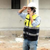 Male architect or engineer using mobile phone for communication while working at construction site. Professional building contractor standing at housing development site uses smartphone on the job. photo