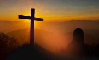 Silhouette of woman holding a bible praying In front of the cross on sunset background, Christian prayer of the Lord photo