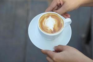 café caliente en manos humanas. flores de espuma de café en una taza blanca. fondo de madera gris foto