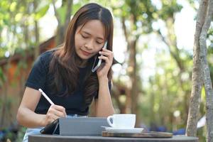 Asian woman looking at earnings on her phone at a coffee shop where she can go to work. amidst green nature photo