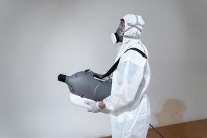 Professional technical man in prevention suit sprays sterilising solution by electrical spray machine on the wood floor and white background with studio light. photo