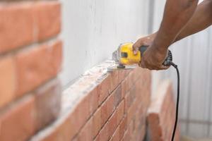 cerca de la máquina trituradora en la mano del hombre trabajador, sostenga la sierra, muela, verifique y mantenga la pared de ladrillo de espuma de poliuretano detallada con polvo por todas partes. foto