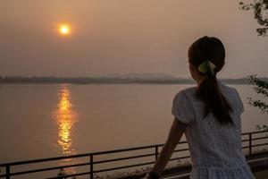 Asian Thai slim woman in traditional cloth stands in front of sunrise time that glitter reflex on Mekong River surface. photo