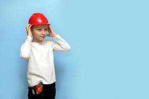 niño con una expresión severa se pone un casco protector de construcción, tiene herramientas en el bolsillo, en un fondo azul con espacio para copiar foto