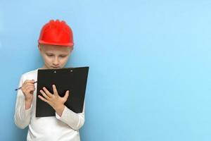 boy in a protective construction helmet concentrating on writing something with a pen on a tablet on a blue background with copy space photo