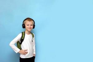 blond boy smiling with a backpack over his shoulders and wearing headphones on a blue background with copy space, back to school photo