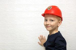 lindo niño rubio sonriente en un casco de seguridad de construcción cerca de una pared blanca con espacio para copiar, orientación profesional foto