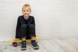 tired smiling cute blond boy sitting on a skateboard after training against a white wall photo
