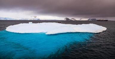 Massive iceberg with blue uderwater part and cruise ship in background, Peterman Island, Antarctica