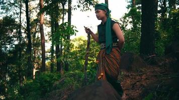 A Man with green clothes and a stick walking through the forest while visiting someone in the village video