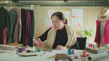Aziatisch middelbare leeftijd vrouw mode ontwerper werken in studio door idee tekening schetsen met digitaal tablet en kleurrijk kleding stof voor een jurk ontwerp verzameling, professioneel winkel kleermaker mkb ondernemer. video