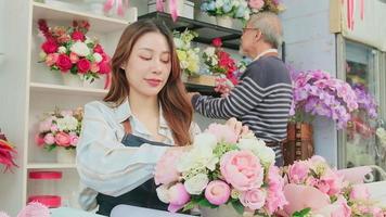 Young beautiful Asian female florist entrepreneur arranging a bunch of blossoms works with old man shopkeeper, happy work in colorful flower shop store with fresh blooms, small business, family SME. video