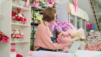One young Caucasian florist owner works with laptop, website arrangement for online business flora service in colorful flower shop store with beautiful bunch of blossom, e-commerce SME technology. video