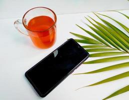 una taza de té, un teléfono de mano y una hoja de palma de fondo blanco aislado en la mañana se ven relajantes foto