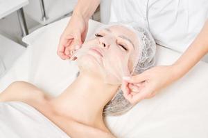 Beautician smoothes sheet mask on woman face for rehydrate face skin, procedure in beauty salon photo