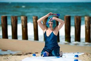 Artistic blue-haired woman performance artist smeared with blue gouache paints sitting on beach photo
