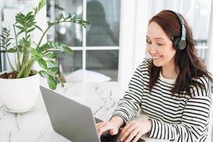 mujer morena sonriente con ropa informal y auriculares que trabajan en una laptop en una oficina moderna y luminosa foto