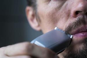 un hombre se corta la barba y el bigote con una maquinilla de afeitar eléctrica, es hora de afeitarse, cuidar la barba y el bigote foto