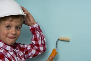 A cute boy in a plaid shirt and a construction helmet with a paint roller in his hands paints a wall photo