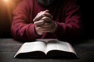 Hands folded in prayer on a Holy Bible in church concept for faith, spirituality and religion, man praying in morning. Man hand with Bible praying. Person Christian who faith in Jesus worship in dark.