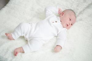 primer plano lindo bebé recién nacido en traje blanco acostado solo en la cama. adorable bebé descansa sobre sábanas blancas, mirando a la cámara con aspecto pacífico. infancia, salud y pediatría, concepto de infancia foto