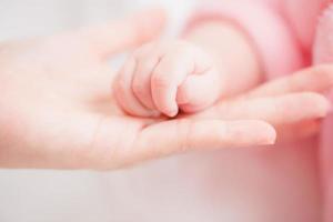 Close up mother holding hands newborn girl in a room. Adorable infant rests on white bedsheets, staring at camera looking peaceful. Infancy, healthcare and paediatrics, babyhood concept. photo