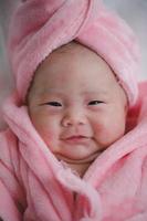 Closeup cute newborn baby in pink bodysuit lying down alone on bed. Adorable infant rests on white bedsheets, staring at camera looking peaceful. Infancy, healthcare and paediatrics, babyhood concept photo