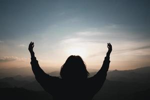 cristianismo de fe del concepto de adoración cristiana. la oración espiritual entrega el brillo del sol con un hermoso fondo de puesta de sol borroso. mujer rezando a dios con bendición esperanzadora contra la puesta de sol. foto