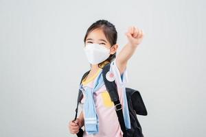 Asian little child girl carry a backpack wearing respirator mask to protect coronavirus outbreak and pointing hand to blank background, New virus Covid-19 photo