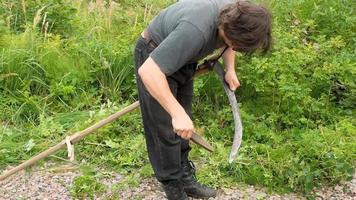 un hombre corta la hierba con una guadaña de mano. cortar la hierba con una guadaña vieja. el agricultor tradicionalmente corta el césped. el cortacésped corta la hierba verde con una guadaña manual de metal. video