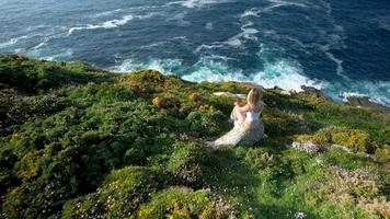 jong vrouw zittend Aan de rots in de heuvels met wilde bloemen en bewonderend de blauw oceaan video