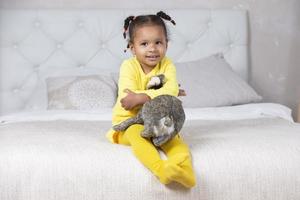 Little African American girl in a yellow dress sits on the bed with a soft toy. photo