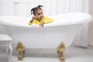 Little African American girl in a yellow dress with curly pigtails sits in a retro bath. photo