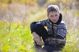 A handsome calm boy in a knitted sweater sits on the grass on the field. photo