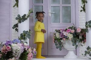 A little African girl in a yellow dress stands on the porch of a house with flowers. photo