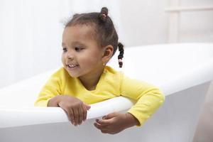 Little African American girl in a yellow dress with curly pigtails sits in a retro bath. photo