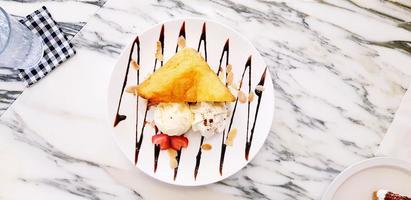 capa plana de tostadas de miel dulce con helado de vainilla, crema batida, jarabe de chocolate y un trozo de fresa en un plato blanco sobre fondo de mármol blanco foto