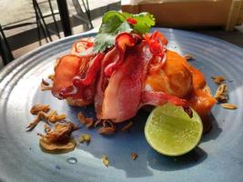 Close up menu of Bangkok eggs Benedict, French toast, grilled bacon with tom yum sauce garnish with cilantro, fried shallots, and a piece of lime, decorated on blue ceramic plate, coffee shop, cafe photo