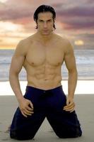 surfista de california muestra su pecho musculoso en la arena mojada de la playa con solo su traje de baño. foto