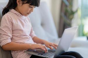 Asian Little school kid girl use laptop computer sitting on sofa alone at home. Child learning reading online social media content, play education lessons game chatting with friends. photo
