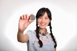 Portrait of beautiful happy young asian woman in denim dress holding crypto currency coin on white background. photo
