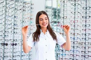 Attractive young female doctor in ophthalmology clinic. Doctor ophthalmologist is standing near shelves with different eyeglasses. photo