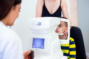 Female oculist using machine for checking eye sight in clinic. Little boy looking at equipment and doctor testing eye pupil in optical store. Concept of eye care and health. photo