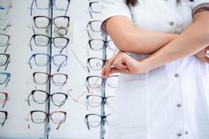 Cropped image of attractive young female doctor in ophthalmology clinic. Doctor ophthalmologist is standing near shelves with different eyeglasses. photo