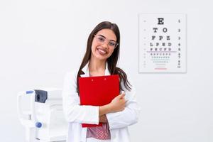 oftalmólogo sonriente sosteniendo portapapeles en el fondo de la tabla optométrica. Docto oculista femenino con un gráfico optométrico detrás de ella foto