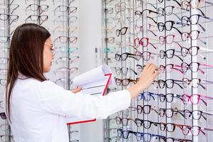 Attractive female ophthalmologist writing data to a clipboard, working in an optical store. Healthcare and medicine concept photo