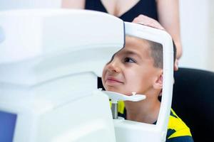 Female oculist using machine for checking eye sight in clinic. Little boy looking at equipment and doctor testing eye pupil in optical store. Concept of eye care and health. photo