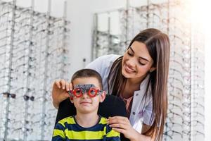 Young smiling boy is sitting and looking at camera through during vision checking. Ophthalmologist is using special medical equipment for eye health saving and improving. photo